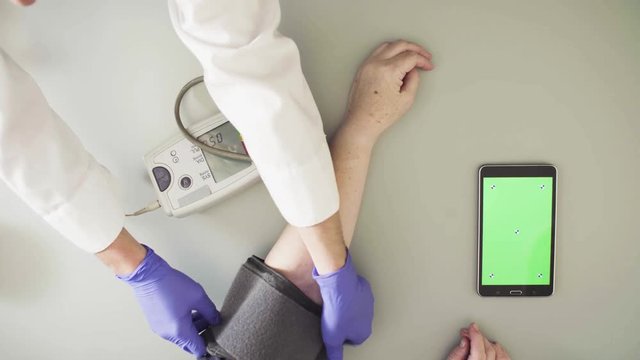 Top view. Doctor measuring blood pressure of senior woman with digital measuring device. A tablet with green screens on the table. Text on the cuff - instructions for use