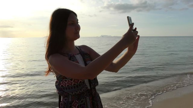 Young beautiful female on beach making selfie during sunset or sunrise