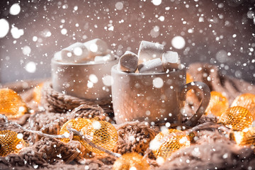 Christmas background with cocoa or coffee with marshmallows in a white cup on a brown knitted winter scarf, snow and a glowing golden garland. Beautiful concept of home comfort and warmth