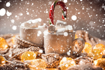 Christmas background with cocoa or coffee with marshmallows and candy in a white cup on a brown knitted winter scarf, snow and a glowing golden garland. Beautiful concept of home comfort and warmth