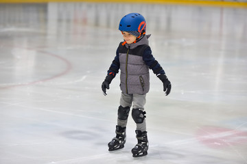 Adorable little boy in winter clothes with protections skating on ice rink