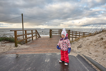 Little girl wear jacket and hat walk is playing at the beach near baltic sea on cloudy sky in winter time