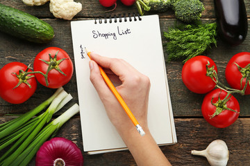 Female hand written shopping list on paper with vegetables on wooden table
