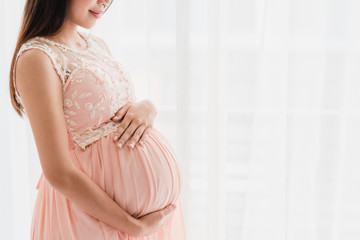 Cropped image of happy beautiful pregnant woman holding pregnant belly near window at home