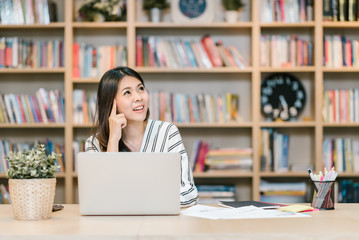 An attractive young Asian woman smiling and thinking of good idea while working at table