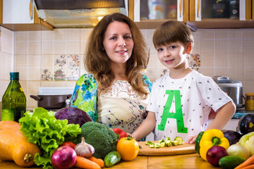 The young cook mother standing with her little son in the kitchen and salting vegetables