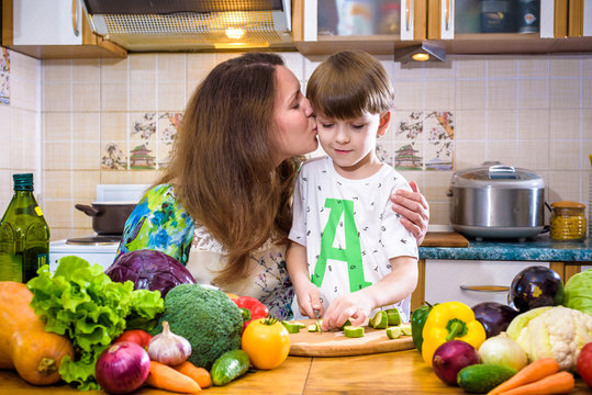 The young cook mother standing with her little son in the kitchen and salting vegetables