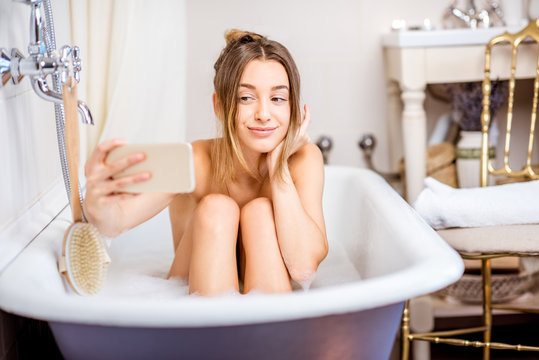 Young and beautiful woman making selfie photo with phone sitting in the retro bath indoors