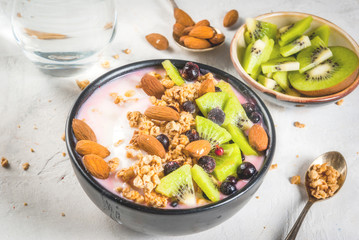 Healthy diet breakfast: smoothies bowl, with yogurt, fresh blueberries (black currant), kiwi, granola oatmeal, almond nuts. On a white stone table, with a glass of water and ingredients. Copy space