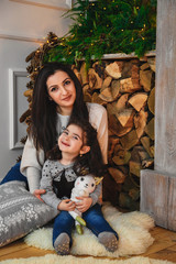 Christmas family portrait of happy smiling mother sitting on the floor near to fireplace decorated with fir and garland and hugging small daughter at home. Winter holiday Xmas and New Year concept