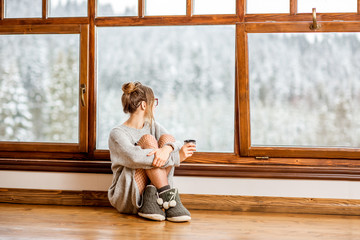 Young woman in sweater sitting near the big window at the cozy wooden mountain house with beautiful landscape view during the winter time