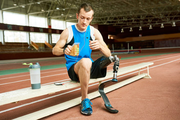 Full length portrait of young amputee athlete taking break from practice on running track and relaxing on bench eating sandwich