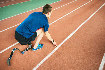 High angle portrait of young amputee athlete on start position on running track in modern indoor stadium, copy space