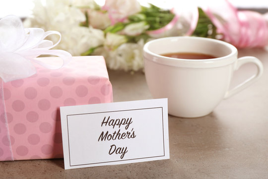 Card with words "Happy Mother's day", gift box and cup of tea on table