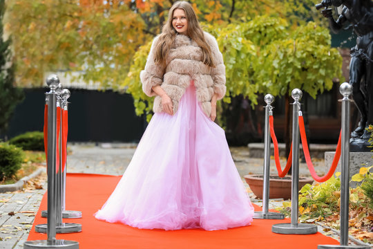 Beautiful young woman in white dress and fur coat on red carpet, outdoors