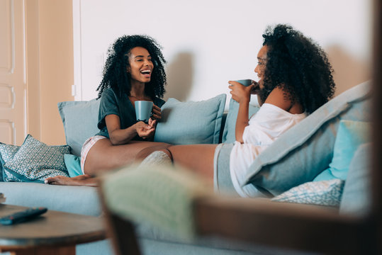 Happy young two black women sitting in the couch drinking coffee .