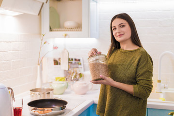 The young cheerful woman cooking in the kitchen and holding dish with buckwheat in hands