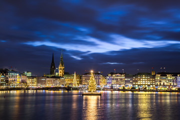 Fototapeta na wymiar Christmas in Hamburg. Panoramic view of the decorated city center from Alster Lake, view to Hamburg Rathaus and a christmas tree installed in the center of the lake. Atmosphere before the New Year. 