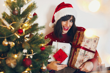 happy stylish woman in santa hat holding many gift boxes with bow at christmas tree lights. space for text. merry christmas and happy new year concept. seasonal greetings, happy holidays