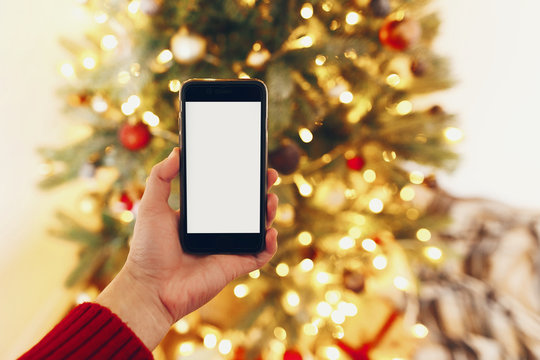 hand holding smart phone with empty screen on background of beautiful christmas tree lights. merry christmas and happy new year concept. mockup, space for text. seasonal greetings, happy holidays