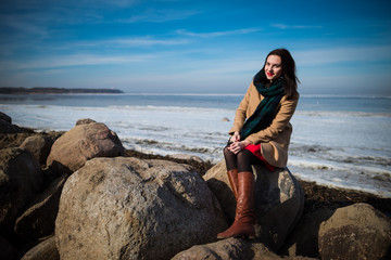 Young beautiful woman in elegant clothes with scarf sits on the coastline in winter season and looking at camera. One person