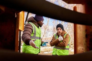 Fototapeta na wymiar Portrait of two workers wearing reflective jackets, one of them African-American, discussing job on industrial site outdoors, shot from inside machine