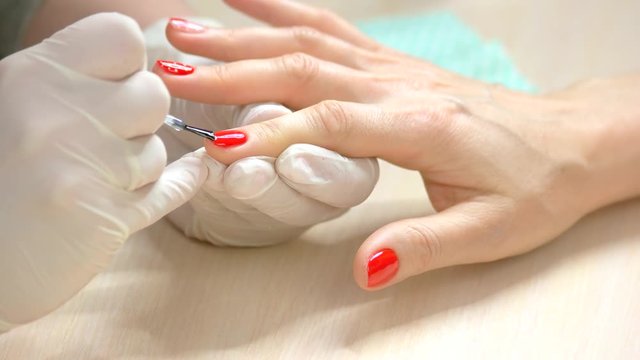 Specialist covers the nail of client by fixer. Nail master fixing red polished index finger nail by transparent fixer in beauty salon close up.
