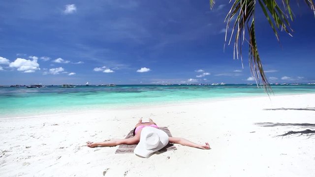 Woman in white straw sunhat lie and relaxing on sandy beach with turquoise sea water and coconut palm trees
