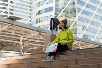 Happy female tourist with suitcase exploring map while sitting in city building, Travel concept