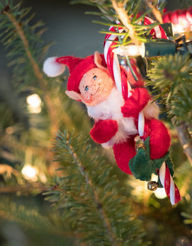 Felt Elf Ornament holding a glass candy cane hanging in a Christmas tree with lights on. Shallow depth of field.
