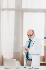 Fototapeta premium portrait of senior doctor in white coat with tablet in hands at workplace in clinic