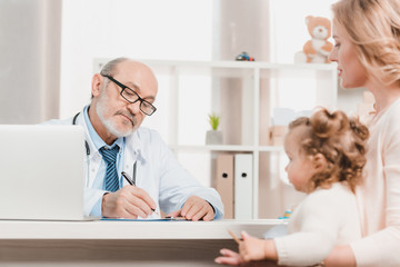 senior doctor making notes while mother and little daughter sitting at table near by in clinic