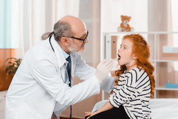 senior pediatrician in medical gloves examining little patients throat in clinic