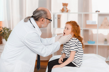 Fototapeta premium senior doctor in medical gloves examining little patients throat in clinic