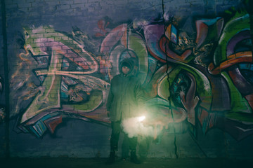 man holding smoke bomb and standing against wall with graffiti at night