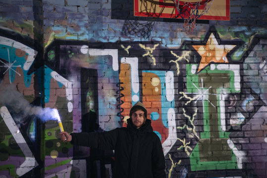 man holding smoke bomb and standing against wall with graffiti at night