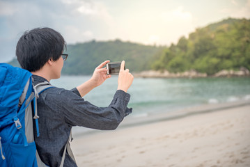 Young Asian backpacker man taking photo of beach and sea by smartphone, summer holiday vacation and travel tropical island concepts