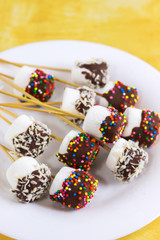 Fototapeta na wymiar marshmallows on a sticks, laying on a white plate. glazed with chocolate and colorful sprinkles.