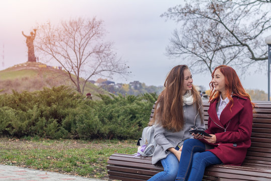  Female teenagers listening to music on smartphone sitting on the bench in an autumn city park