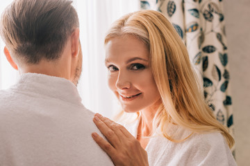 Obraz na płótnie Canvas beautiful mature woman smiling at camera while embracing husband in bathrobe in hotel room
