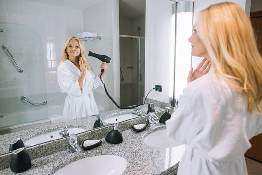 attractive mature woman in bathrobe drying hair with hair dryer in bathroom at hotel