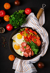 Breakfast. Fried eggs with sausage and vegetables in a frying pan on a black background in rustic style. Top view