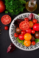 Tomatoes of different colors with green herbs in a bowl on a black background. Flat lay. Top view