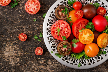 Tomatoes of different colors with green herbs in a bowl on a black background. Flat lay. Top view