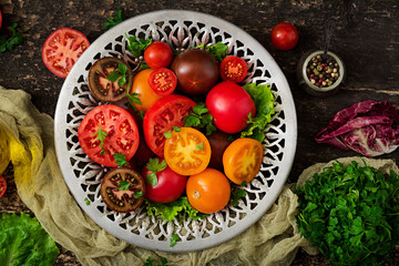 Tomatoes of different colors with green herbs in a bowl on a black background. Flat lay. Top view