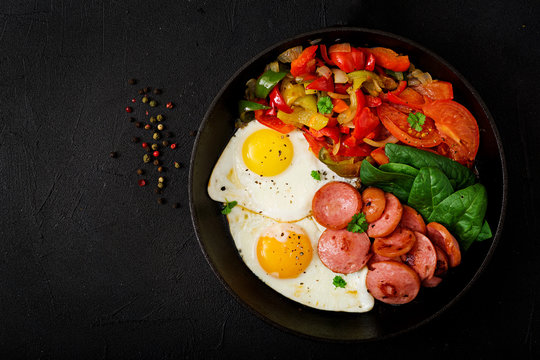 Breakfast. Fried eggs with sausage and vegetables in a frying pan on a black background in rustic style. Top view