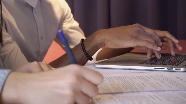 Close-up African American businessman of an international team seriously working at one laptop. Multi-ethnic team running on desktop with papers and computer, contract, another employee draws diagram.