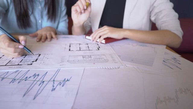 Two female realtors prepares for the presentation sitting in the office. The papers with the house schemes and market prices charts lie on the big wooden table and professional outlines the important