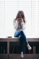 Woman drinking hot coffee sitting on window sill at home