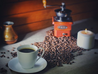 coffee beans on a wooden table, next to a cup of hot coffee, a hand grinder and a candle